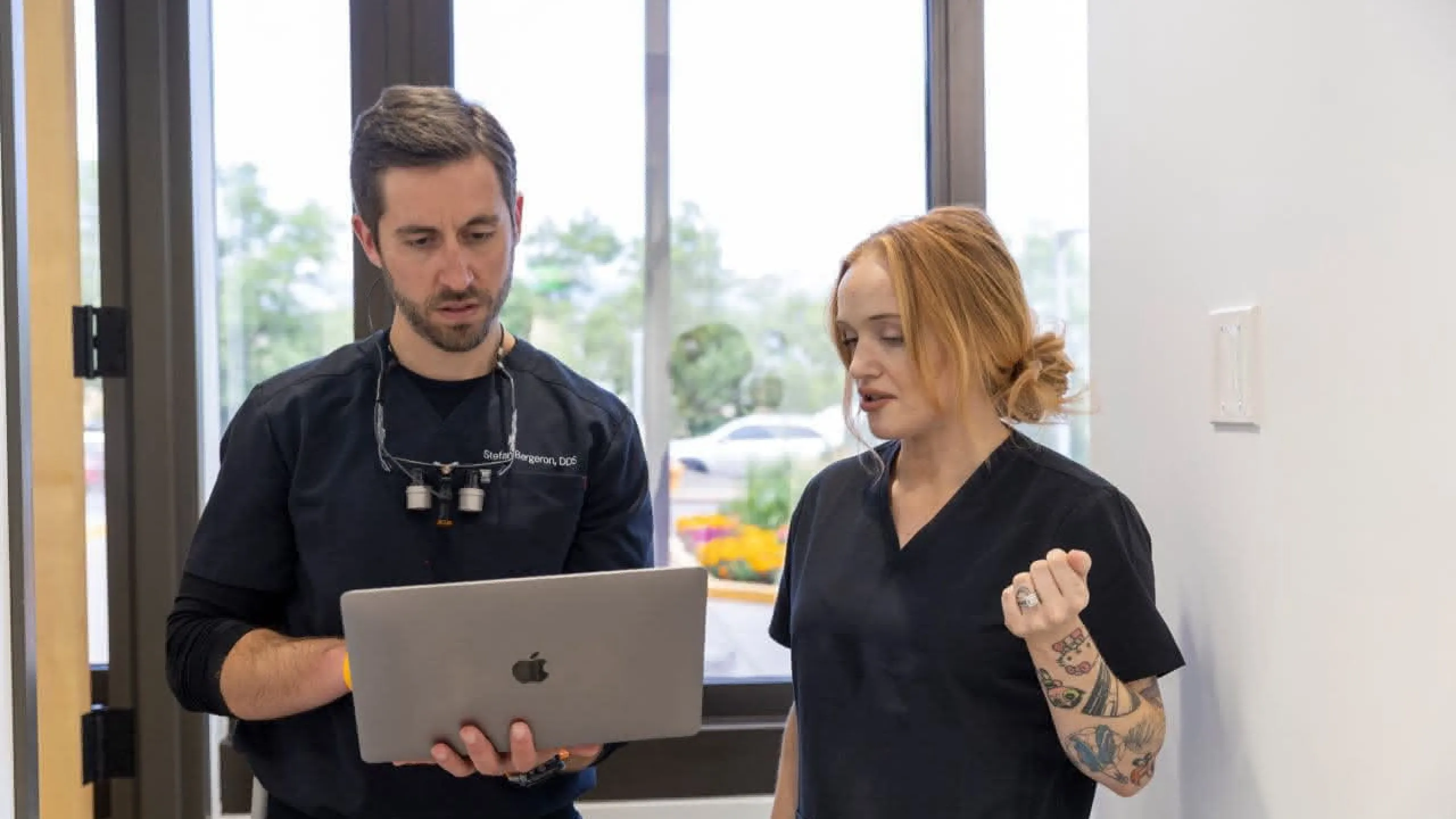 A dentist holding a laptop with a dental assistant in the hallway