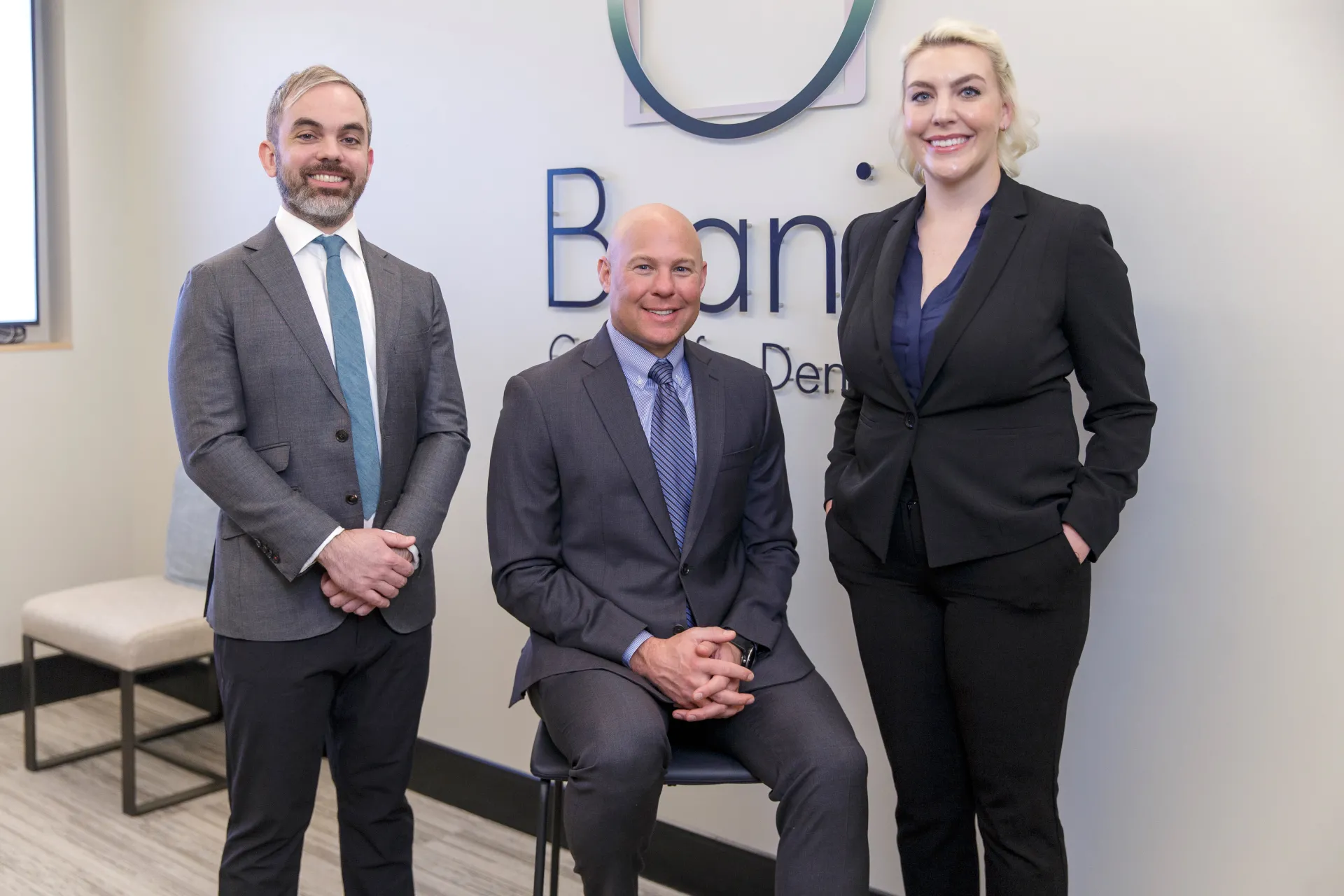 Dr. John Sporidis, Dr. Scott Dudley, and Dr. Claire Sesson smiling together in front of the Branin Center for Dentistry Logo